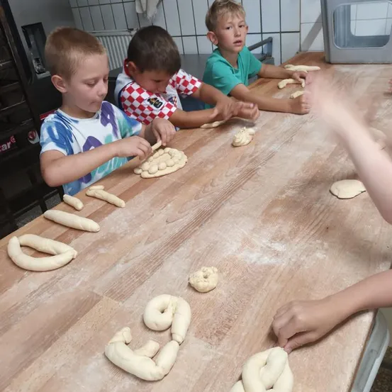 Drei Jungen sitzen an einem Holztisch und beschäftigen sich mit dem Formen von Teig. Der Tisch ist mit Teig und Mehl bedeckt. Ein Junge formt die Zahl 39 aus Teig.