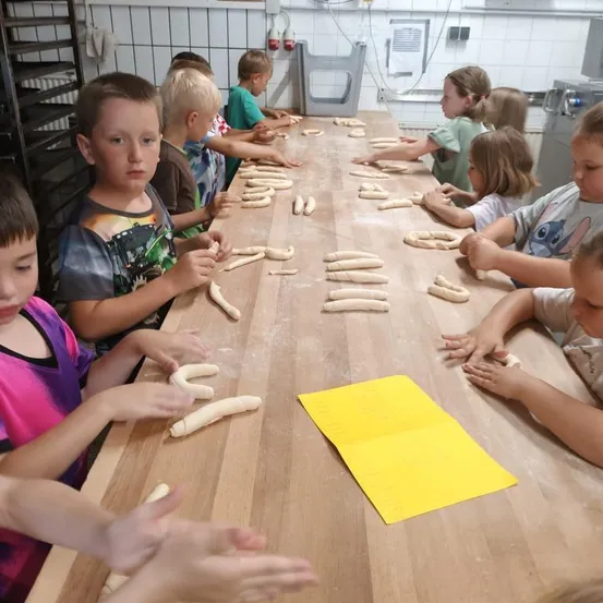 Eine Gruppe von Kindern sitzt um einen langen Holztisch herum, wobei jedes Kind Formen aus Teig macht. Der Tisch ist mit Teig bedeckt und ein gelbes Papier liegt in der Mitte.