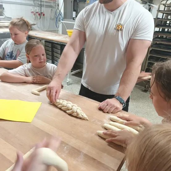 Ein Mann in einem weißen T-Shirt mit Logo unterrichtet Kinder, wie man Teig auf einem Holztisch flicht.