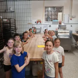 Eine Gruppe von Kindern steht um einen langen Tisch in einer Küche herum, lächelt und posiert für ein Foto. Ein Mann steht in der Nähe einer Maschine im Hintergrund.