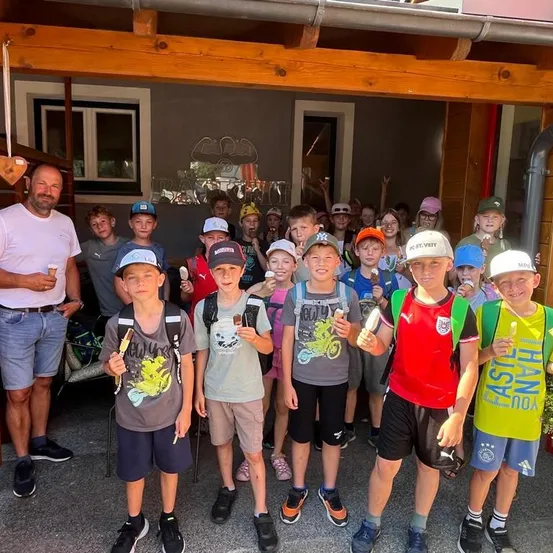 Eine Gruppe von Kindern und einem Mann stehen vor einem Gebäude und halten Eis am Stiel. Die Kinder tragen Hüte und Turnschuhe, und der Mann trägt ein weißes Hemd und Jeans.