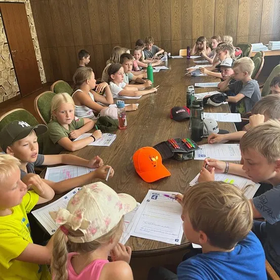 Eine Gruppe von Kindern sitzt um einen langen Tisch herum und ist in einer Klassenzimmeraktivität engagiert. Papiere, Stifte und Mützen sind auf dem Tisch verstreut. Einige Kinder schauen auf ihre Papiere, während andere umherschauen.
