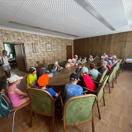 Eine Gruppe von Kindern und Erwachsenen sitzt um einen großen Tisch in einem Raum mit Holzwänden und einem gemusterten Decken. Einige tragen Hüte und Brillen. Eine Frau steht in der Nähe der Tür.