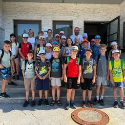 Eine Gruppe von Kindern und Erwachsenen steht auf den Stufen eines Gebäudes, alle tragen Mützen und haben Rucksäcke dabei. Sie lächeln und posieren für ein Foto.