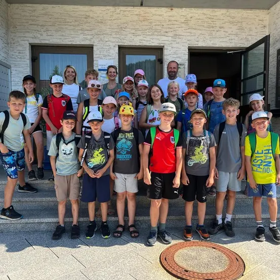 Eine Gruppe von Kindern und Erwachsenen steht auf den Stufen eines Gebäudes, alle tragen Mützen und haben Rucksäcke dabei. Sie lächeln und posieren für ein Foto.