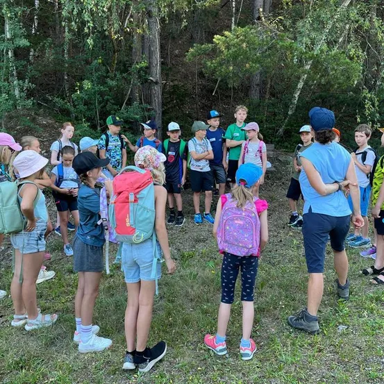 Eine Gruppe von Kindern und Erwachsenen steht in einem Kreis in einem Waldgebiet, einige tragen Hüte und haben Rucksäcke dabei. Eine Frau in einem blauen Shirt steht vor ihnen.