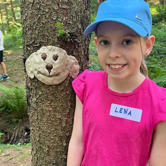 Ein junges Mädchen namens Lena steht neben einem Baum mit einem geschnitzten Hundegesicht. Sie lächelt und trägt ein rosa Shirt und eine blaue Kappe.