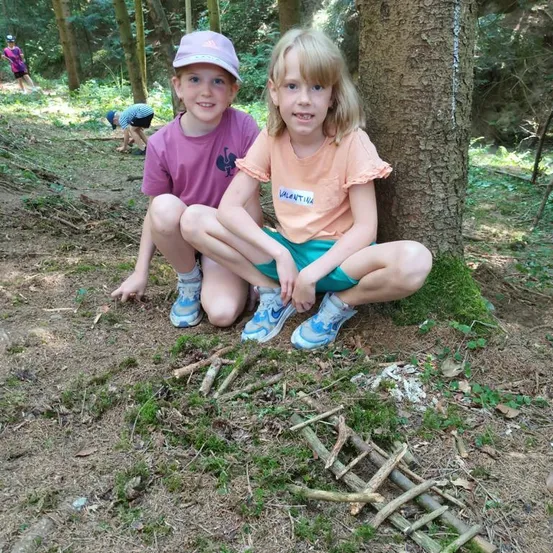 Zwei junge Mädchen sitzen auf dem Boden in einem Wald, eines trägt ein lila Shirt und das andere ein orangefarbenes Shirt mit Namensschild. Sie lächeln und posieren für ein Foto.