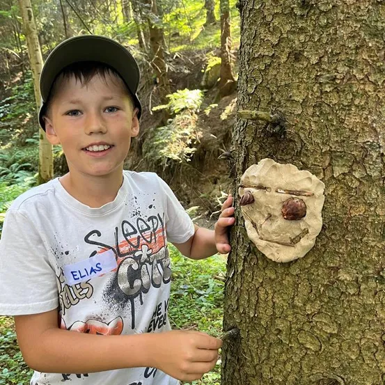 Ein junger Junge im Wald, der lächelt und einen Baum mit einem Gesicht aus Rinde und Zweigen berührt.