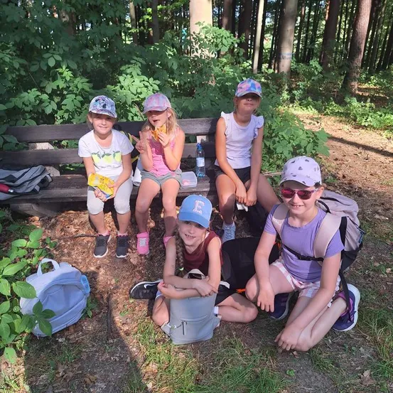 Fünf junge Mädchen sitzen auf einer Bank im Wald, lächeln und posieren für ein Foto. Sie tragen Hüte und Turnschuhe, und einige von ihnen halten Snacks.