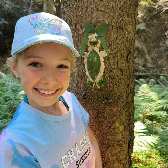 Ein junges Mädchen mit einer Schmetterlingsmütze lächelt neben einem Baum mit einer aus Blättern gefertigten Schildkröte.