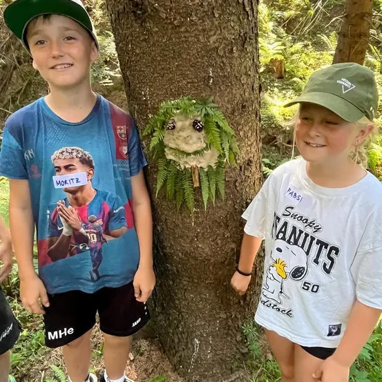Zwei Kinder stehen neben einem Baum in einem Wald, eines trägt ein Shirt mit einem Fußballspieler und das andere ein Shirt mit Snoopy.