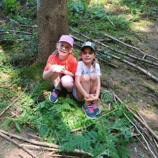 Zwei junge Mädchen sitzen auf dem Boden in einem Wald, eines trägt einen rosa Hut und das andere eine grüne Mütze. Sie sind von Farnen und Stöcken umgeben, mit einem Baumstamm hinter ihnen.