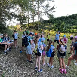 Eine Gruppe von Kindern und Erwachsenen in einem Wald, einige stehend und einige sitzend, tragen Hüte und tragen Rucksäcke, mit einer Frau in einem blauen Shirt, die sie führt.