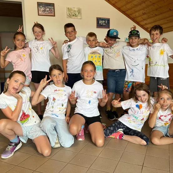 Eine Gruppe von Kindern in einem Raum, einige sitzend und einige stehend, alle tragen weiße T-Shirts mit Zeichnungen und lächeln für ein Foto.