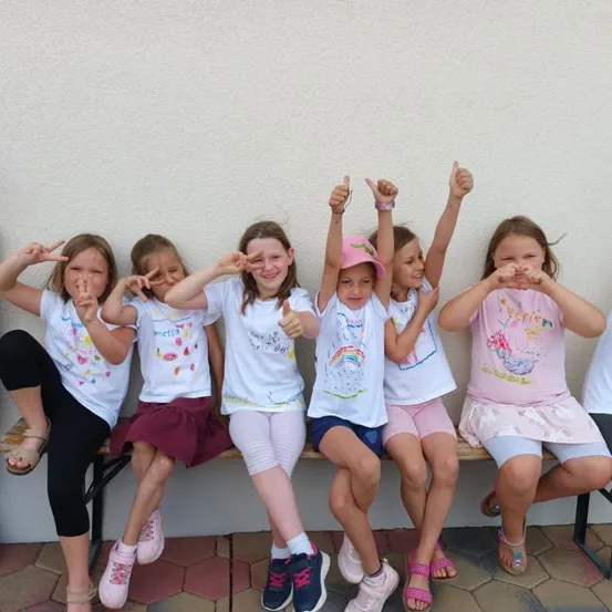 Eine Gruppe junger Mädchen in bunten T-Shirts und Röcken sitzt auf einer Bank, lächelt und macht Friedenszeichen. Sie posieren für ein Foto.