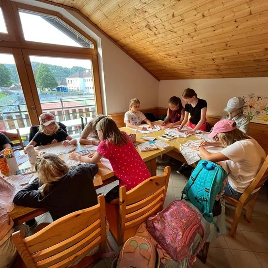 Eine Gruppe von Kindern sitzt um einen Tisch in einem Klassenzimmer und ist mit einer Aktivität beschäftigt. Einige malen, während andere zeichnen. Der Raum hat Holzwände und ein Fenster mit Blick aufs Freie.