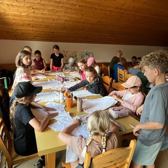 Eine Gruppe von Kindern und Jugendlichen sitzt um einen Tisch herum und ist mit einer Bastelaktivität beschäftigt. Sie sitzen auf Stühlen, einige stehen. Der Tisch ist mit weißen Tüchern und verschiedenen Bastelmaterialien bedeckt. Der Raum hat Holzwände und eine Holzdecke.
