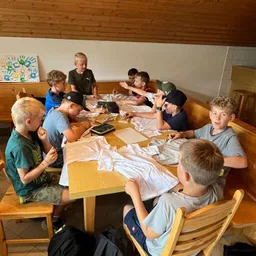 Eine Gruppe von Jungen sitzt um einen Holztisch herum und ist mit einer Bastelaktivität beschäftigt. Sie tragen Freizeitkleidung und einige tragen Mützen. Der Tisch ist mit weißem Tuch bedeckt und hat verschiedene Gegenstände darauf. Der Raum hat Holzwände und eine weiße Wand mit einem farbenfrohen Handabdruck.