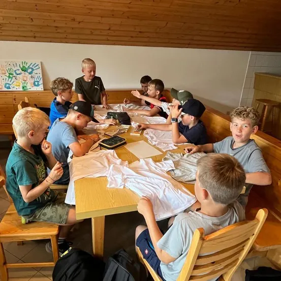 Eine Gruppe von Jungen sitzt um einen Holztisch herum und ist mit einer Bastelaktivität beschäftigt. Sie tragen Freizeitkleidung und einige tragen Mützen. Der Tisch ist mit weißem Tuch bedeckt und hat verschiedene Gegenstände darauf. Der Raum hat Holzwände und eine weiße Wand mit einem farbenfrohen Handabdruck.