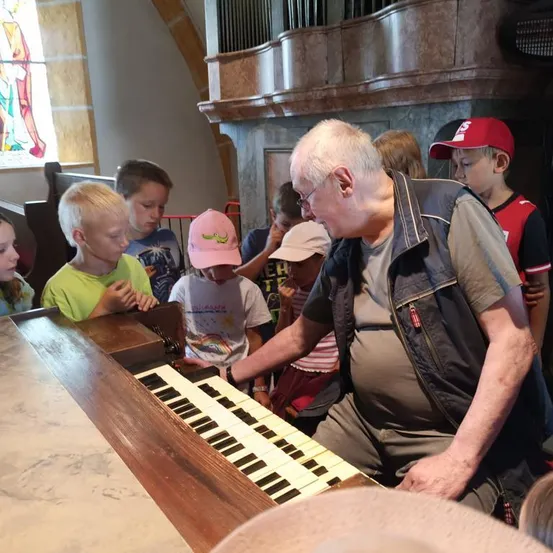 Ein älterer Mann spielt Orgel in einer Kirche, während mehrere Kinder aufmerksam zuschauen.