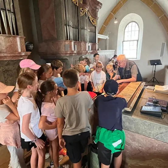 Eine Gruppe von Kindern und ein älterer Mann versammeln sich um ein Klavier in einer Kirche, mit einer großen Orgel hinter ihnen.