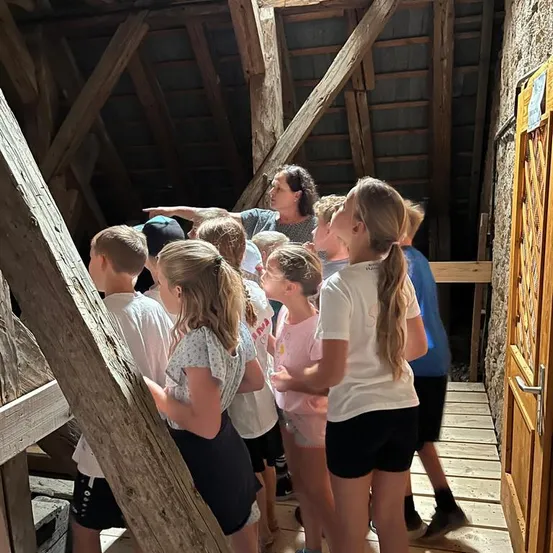Eine Gruppe von Kindern und einer erwachsenen Frau versammelt sich in einer Scheune, wobei die Frau auf etwas über ihnen zeigt. Die Kinder schauen auf und scheinen an der Aktivität interessiert zu sein.