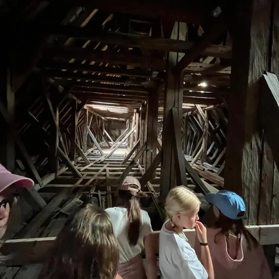 Eine Gruppe von Menschen erkundet das Innere einer alten Holzstruktur, möglicherweise ein Scheune oder Silo. Sie stehen auf Holzplanken und schauen nach oben zu den Holzbalken.