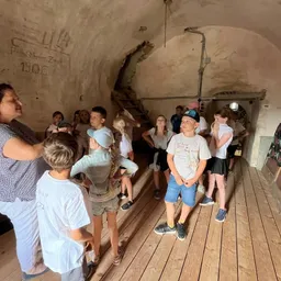 Eine Gruppe von Kindern und einer erwachsenen Frau versammelt sich in einem schwach beleuchteten Raum mit Holzböden und lauscht einer sprechenden Frau. Der Raum hat eine Treppe und Rohre an den Wänden.