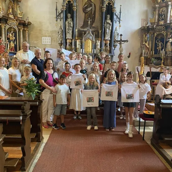 Eine Gruppe von Kindern und Erwachsenen steht in einer Kirche und hält weiße Papiere mit Zeichnungen hoch. Sie lächeln und posieren für ein Foto.
