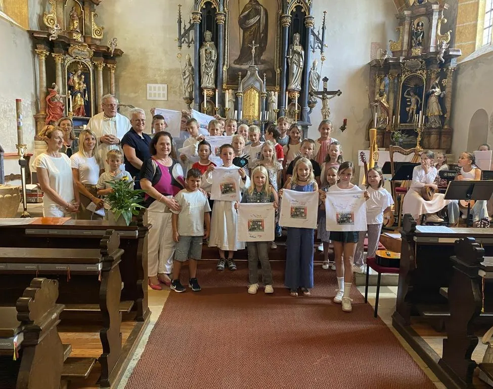 Eine Gruppe von Kindern und Erwachsenen steht in einer Kirche und hält weiße Papiere mit Zeichnungen hoch. Sie lächeln und posieren für ein Foto.