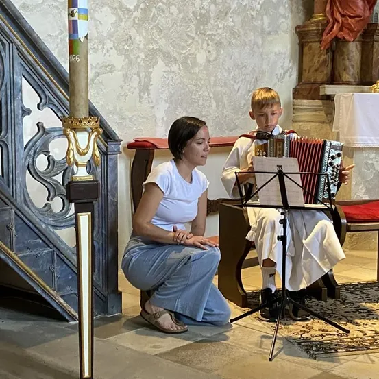 Eine Frau kniet neben einem Jungen, der in einer Kirche ein Akkordeon spielt, beide sehen sich Noten auf einem Ständer an.