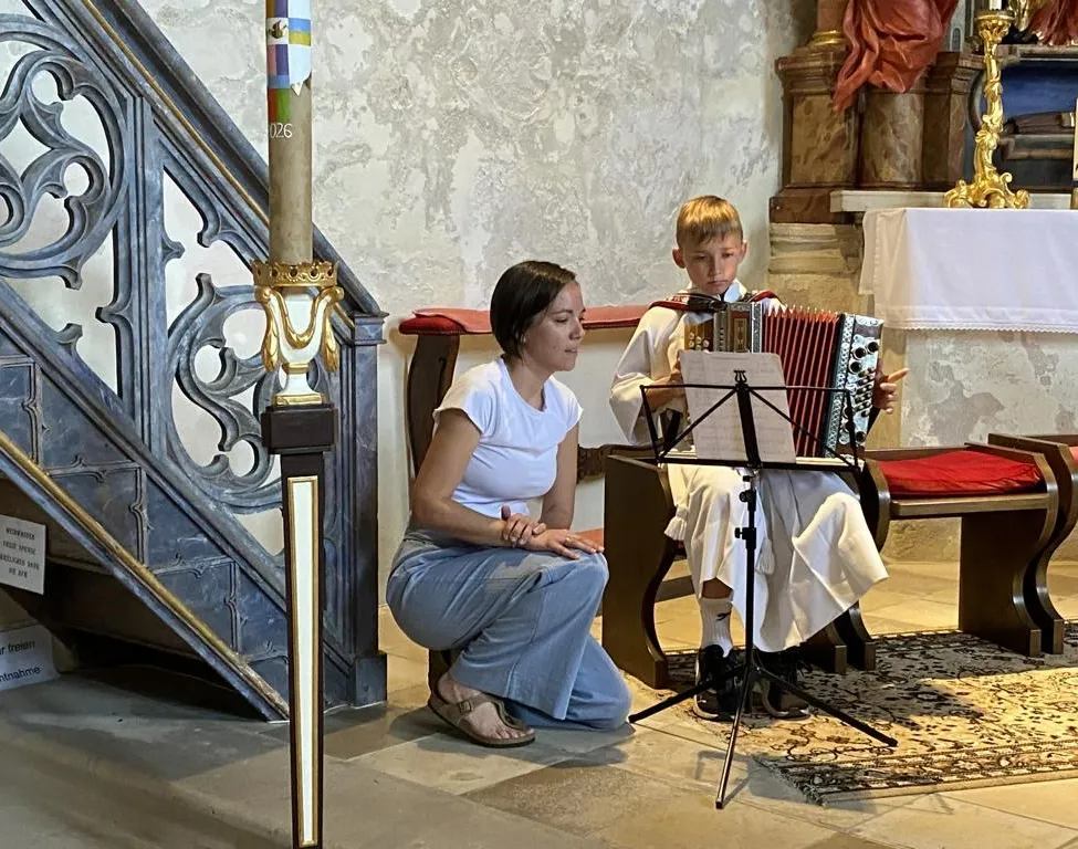 Eine Frau kniet neben einem Jungen, der in einer Kirche ein Akkordeon spielt, beide sehen sich Noten auf einem Ständer an.