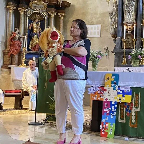 Eine Frau steht in einer Kirche, hält eine Puppe in einer Hand und ein Mikrofon in der anderen, während ein Priester hinter ihr sitzt. Ein farbenfrohes Puzzle-Kreuz steht auf dem Altar.