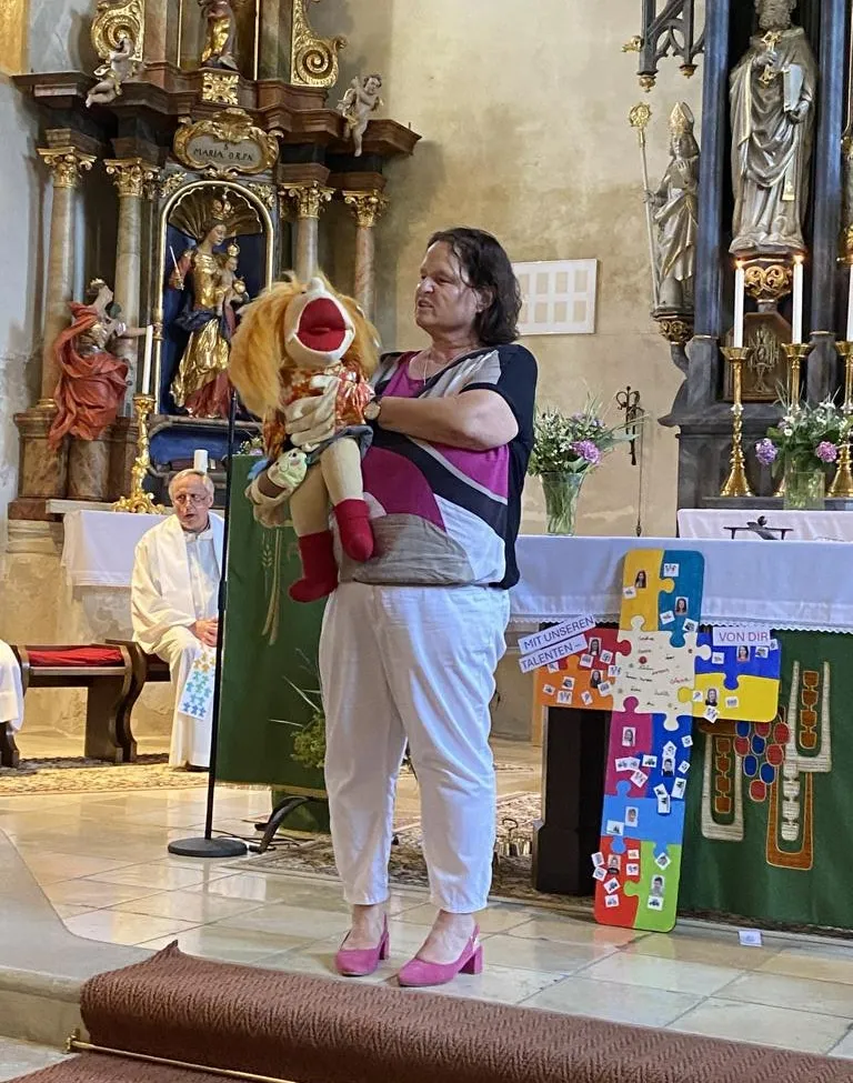 Eine Frau steht in einer Kirche, hält eine Puppe in einer Hand und ein Mikrofon in der anderen, während ein Priester hinter ihr sitzt. Ein farbenfrohes Puzzle-Kreuz steht auf dem Altar.