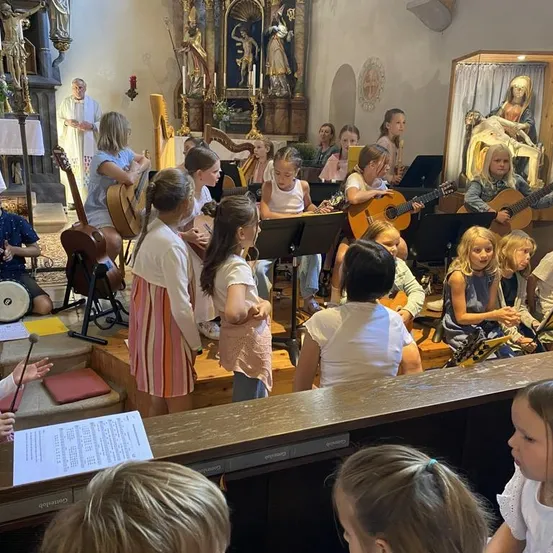 Eine Gruppe von Kindern spielt verschiedene Musikinstrumente in einer Kirche. Einige spielen Gitarren, während andere Flöten und Trommeln spielen. Ein Priester steht hinter ihnen.