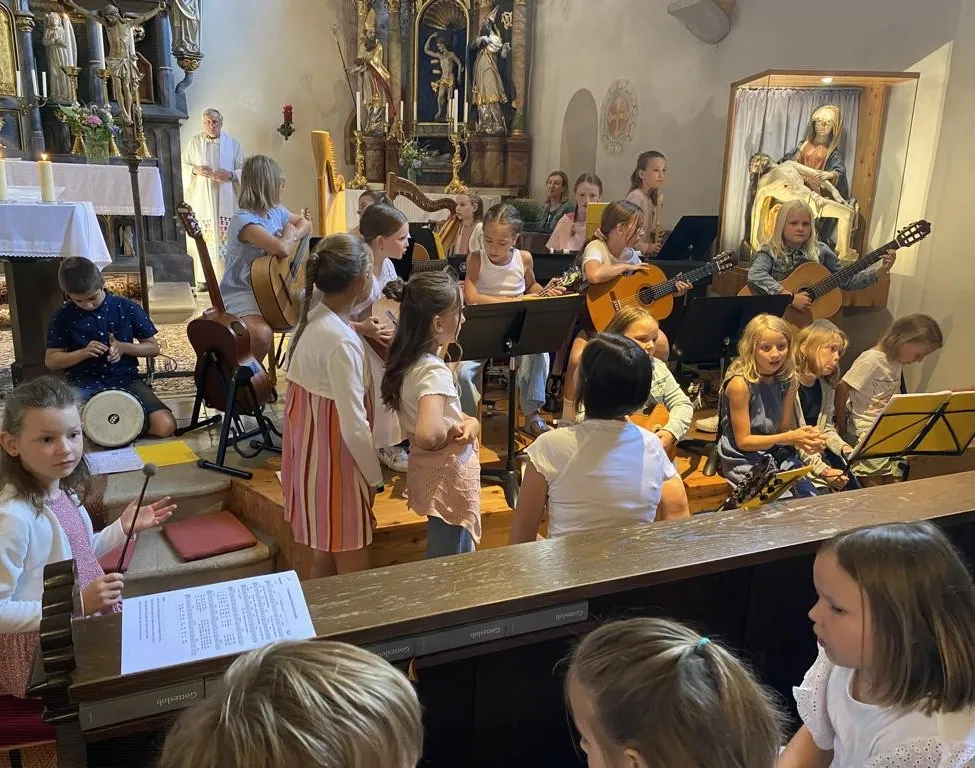 Eine Gruppe von Kindern spielt verschiedene Musikinstrumente in einer Kirche. Einige spielen Gitarren, während andere Flöten und Trommeln spielen. Ein Priester steht hinter ihnen.