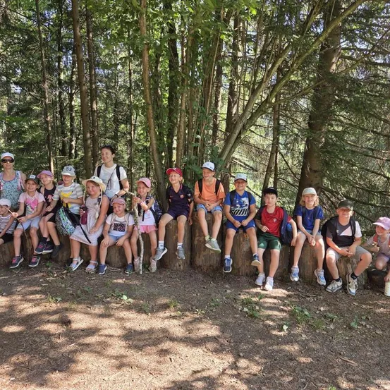 Eine Gruppe von Kindern und Erwachsenen posiert für ein Foto im Wald, auf Baumstümpfen sitzend. Sie tragen alle Mützen und Turnschuhe.