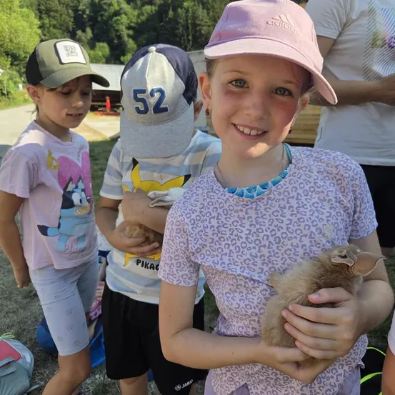 Drei Kinder im Freien, eines hält ein kleines Kaninchen, ein anderes ein kleines Tier und ein drittes schaut nach unten.
