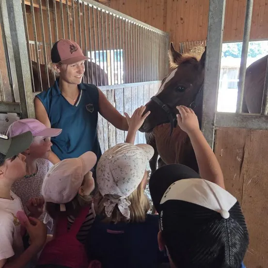 Eine Frau in einer blauen Weste steht in einem Stall mit einem Pferd, umgeben von Kindern, die das Pferd streicheln.