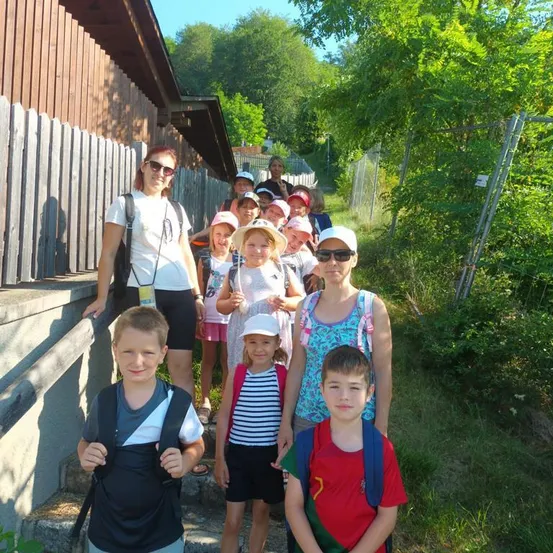 Eine Gruppe von Kindern und Erwachsenen steht auf einem Pfad, wahrscheinlich auf einer Schulfahrt. Sie tragen alle Hüte und haben Rucksäcke dabei. Die Frau vorne trägt eine Sonnenbrille und ein weißes T-Shirt. Hinter ihnen steht ein Holzzaun und ein Gebäude.