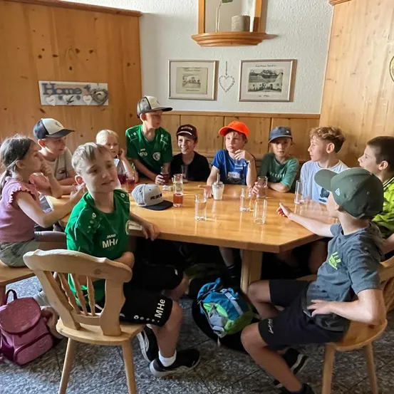 Eine Gruppe von Kindern in einem Raum mit Holzwänden, die um einen Tisch mit Gläsern und Strohhalmen sitzen, einige tragen Mützen und Turnschuhe.