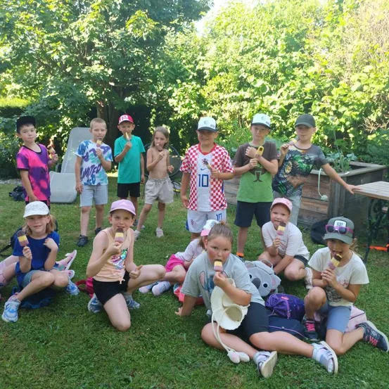 Eine Gruppe von Kindern in einem Garten, einige stehend und einige sitzend, alle mit Eis am Stiel. Sie sind von Grünpflanzen und einem Holztisch umgeben.
