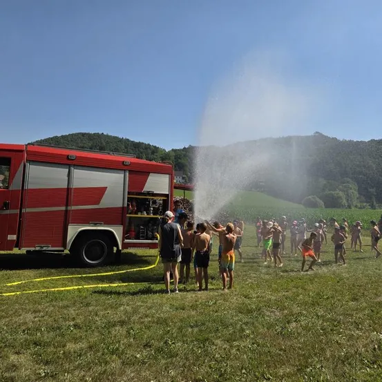 Eine Gruppe von Menschen spielt an einem sonnigen Tag mit dem Wasserschlauch eines Feuerwehrautos.