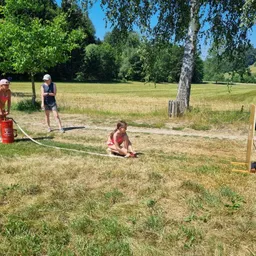 Drei Personen spielen ein Spiel auf einem Feld. Ein Mädchen kniet mit einem roten Objekt, während zwei Frauen in der Nähe stehen. Bäume und ein Feuerlöscher sind sichtbar.