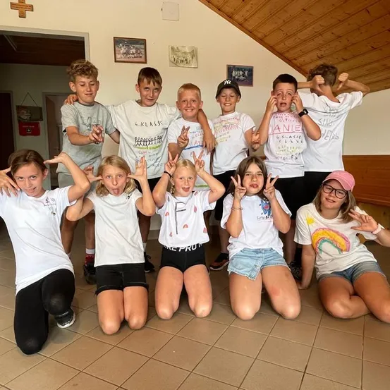 Eine Gruppe von Kindern in weißen T-Shirts posiert für ein Foto in einem Raum mit Holzbalken und gefliestem Boden. Einige machen Friedenszeichen und andere haben die Hände hoch.