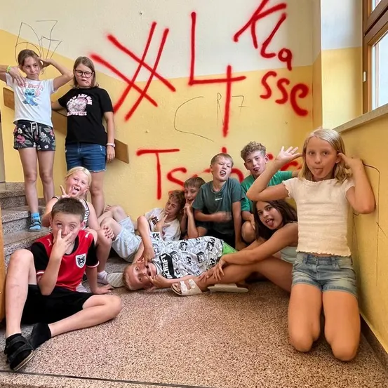 Eine Gruppe von Kindern und Teenagern posiert für ein Foto in einem Flur mit Graffiti an der Wand. Einige sitzen auf dem Boden, während andere auf den Stufen stehen.