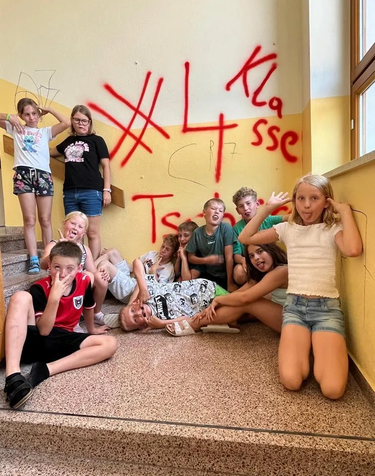 Eine Gruppe von Kindern und Teenagern posiert für ein Foto in einem Flur mit Graffiti an der Wand. Einige sitzen auf dem Boden, während andere auf den Stufen stehen.