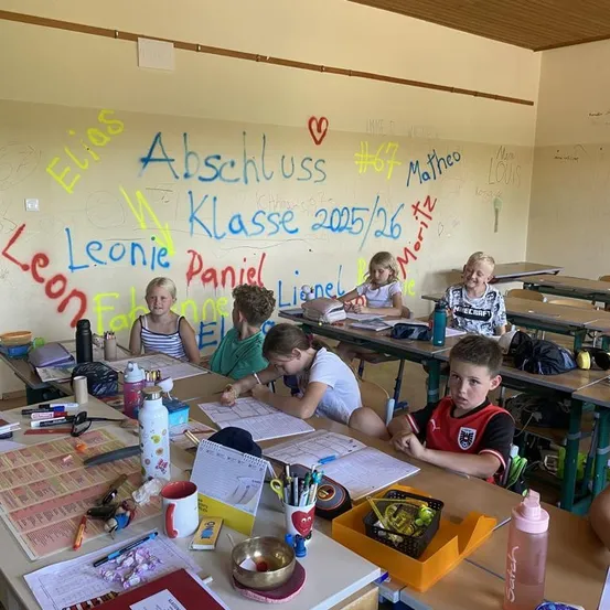 Ein Klassenzimmer mit Schülern an Schreibtischen, die schreiben. An der Wand dahinter befinden sich bunte Graffiti, einschließlich Namen und eines Herzsymbols. Verschiedene Schulmaterialien sind auf den Tischen verstreut.