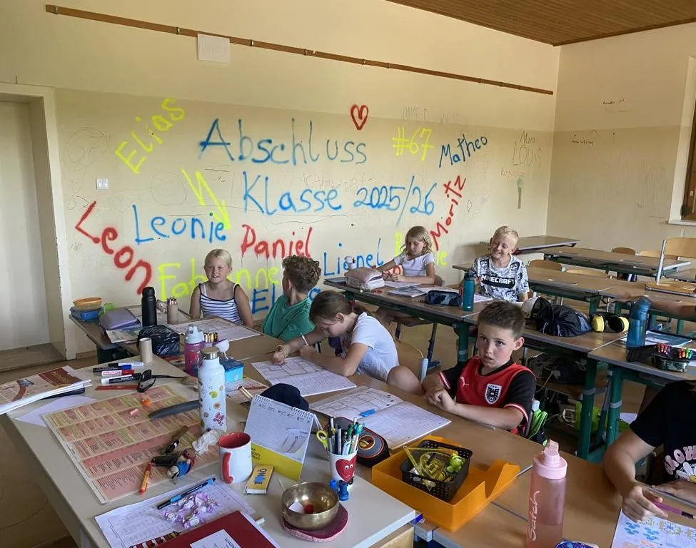 Ein Klassenzimmer mit Schülern an Schreibtischen, die schreiben. An der Wand dahinter befinden sich bunte Graffiti, einschließlich Namen und eines Herzsymbols. Verschiedene Schulmaterialien sind auf den Tischen verstreut.